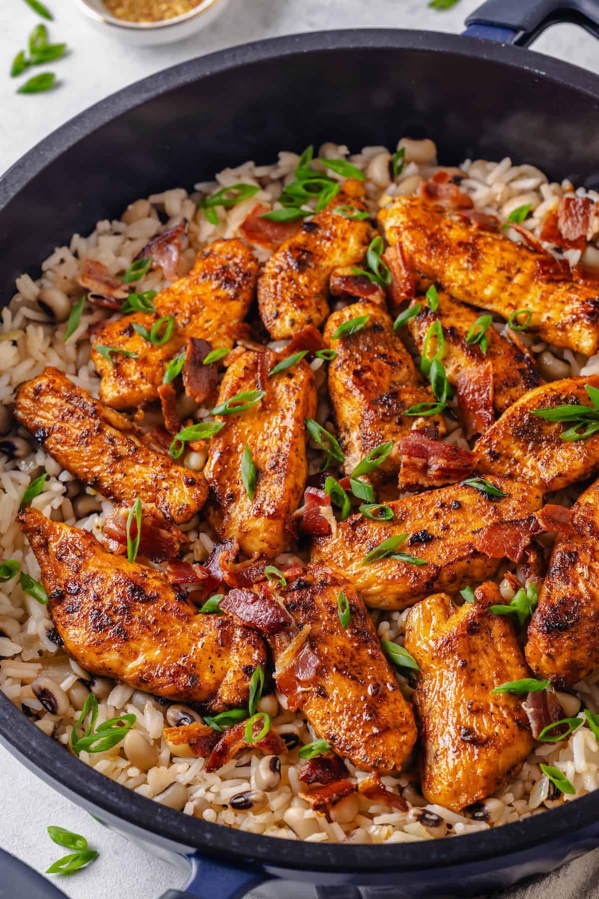 Spiced chicken strips served atop a pot of rice with black eyed peas topped with sliced green onions and cooked, crumbled bacon.