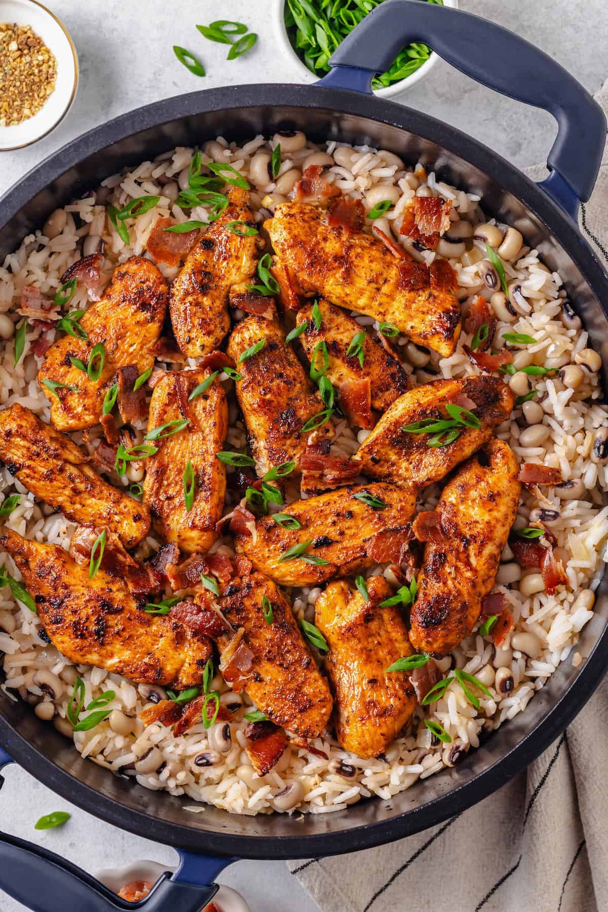 Spiced chicken strips on top of a pot of rice with black eyed peas topped with sliced green onions and cooked, crumbled bacon.