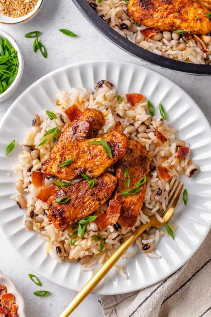 A white plate with a serving of rice and black eyed peas with spiced chicken on top and a fork to the side.