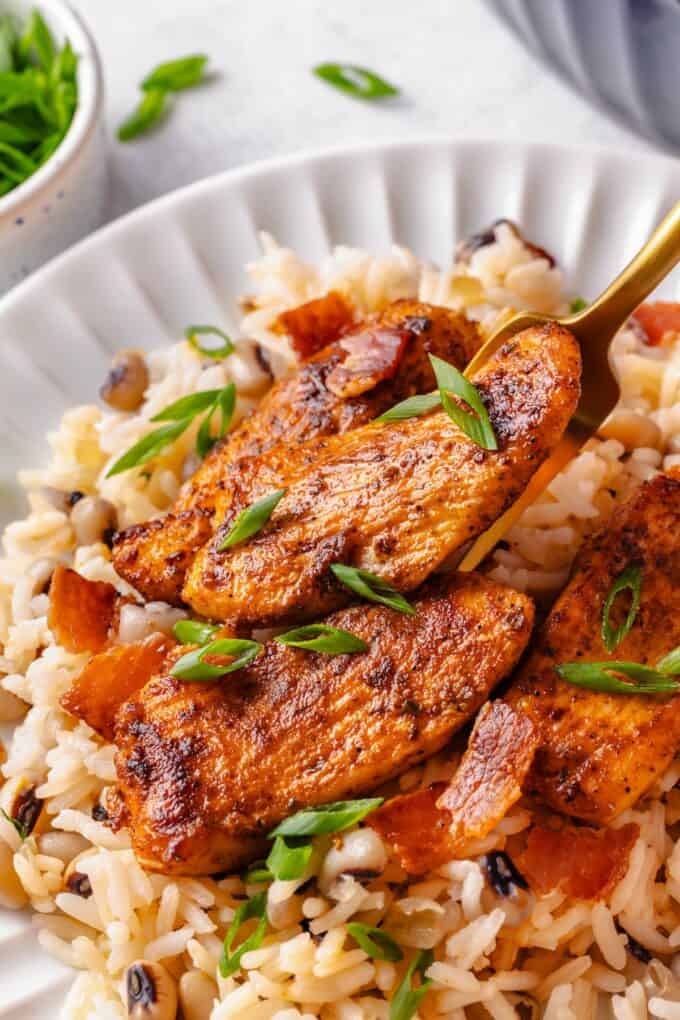 A fork lifting up a seasoned strip of chicken breast over a bed of rice with black eyed peas on a white plate.