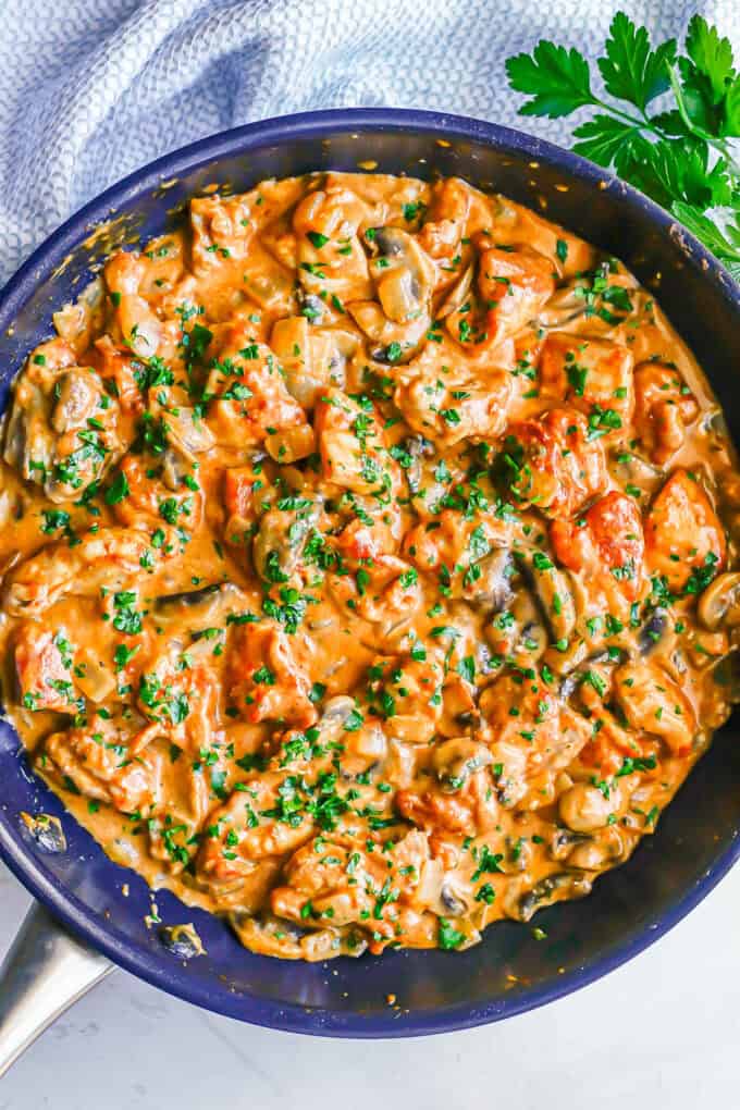 A large dark skillet with a creamy paprikash chicken mixture with onions and mushrooms and chopped fresh parsley on the top.