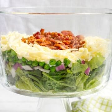 A wide glass bowl with a layered salad of spinach, red onion, peas, cheese, bacon and a creamy dressing.