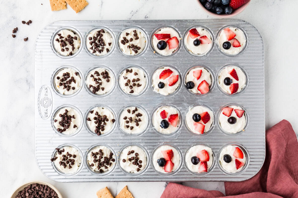 A mini muffin tin with individual cheesecake bites, half topped with mini chocolate chips and half topped with blueberries and cut strawberries.
