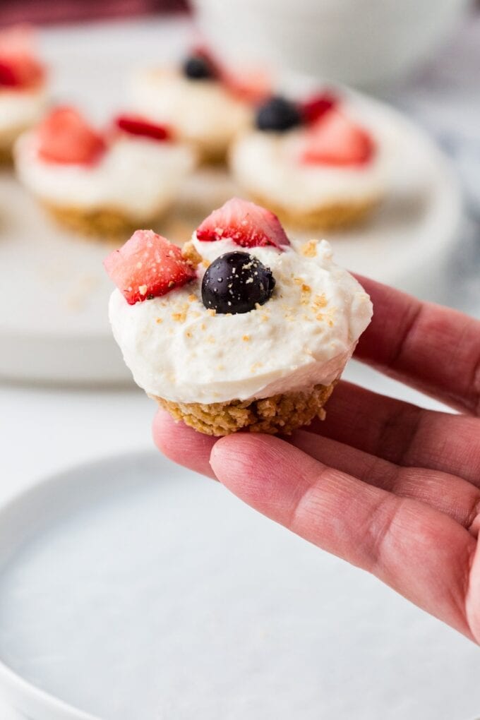 A hand holding up an individual graham cracker crust mini cheesecake with fresh berries on top.