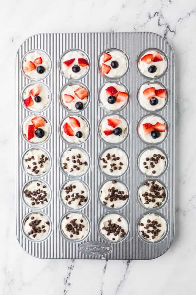 A mini muffin tin with mini cheesecake bites, half topped with fresh berries and half topped with mini chocolate chips.