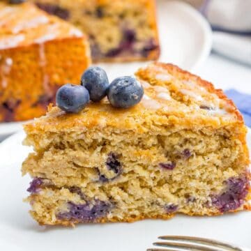 Close up of a slice of homemade blueberry cake with a light lemon glaze and fresh blueberries on top on a white plate.