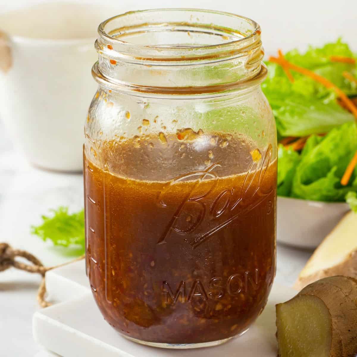 Close up of a glass jar with a homemade soy ginger vinaigrette with sliced ginger pieces to the side.