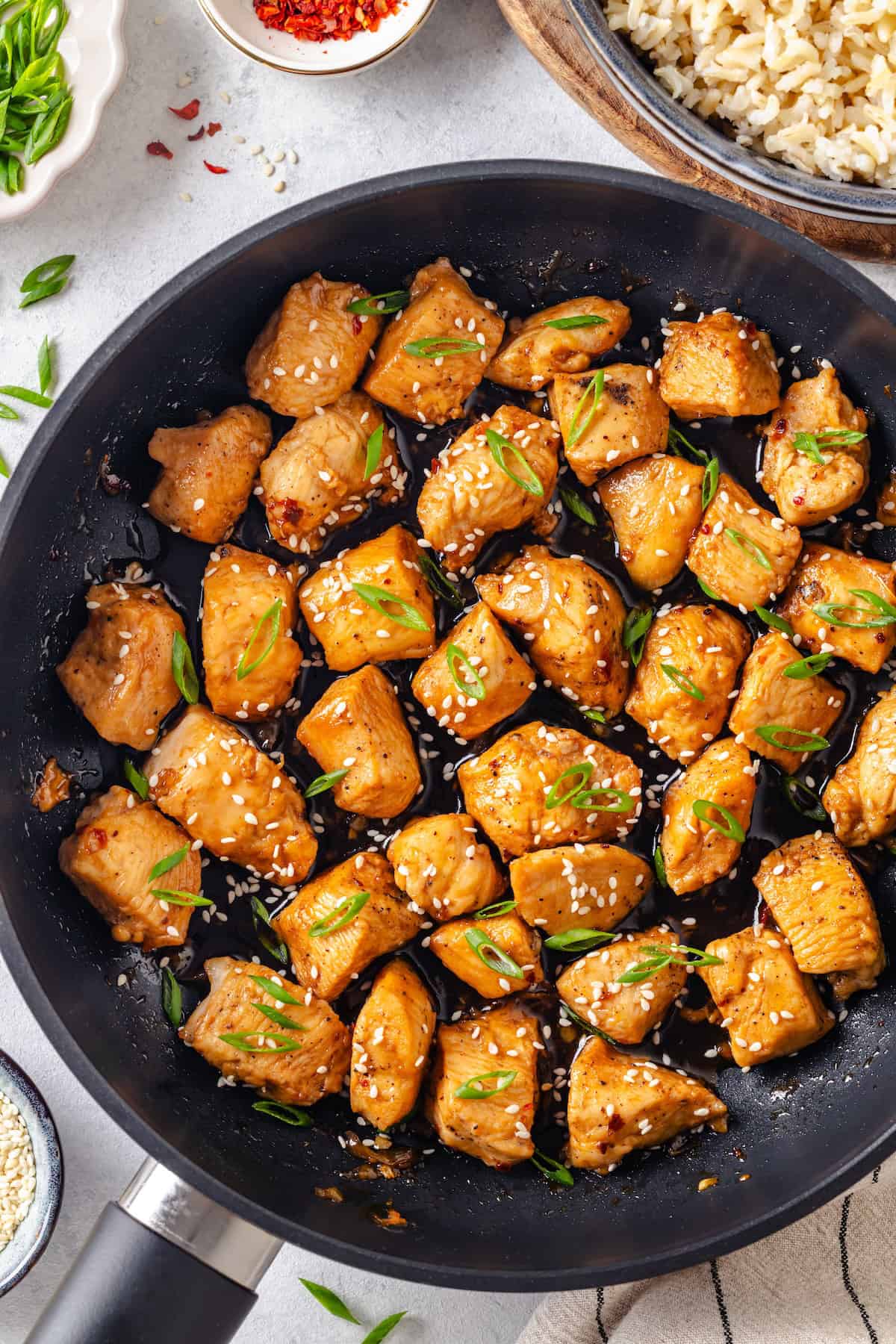 A saucy honey garlic chicken skillet with sesame seeds and sliced green onions sprinkled on top and a bowl of rice to the side.