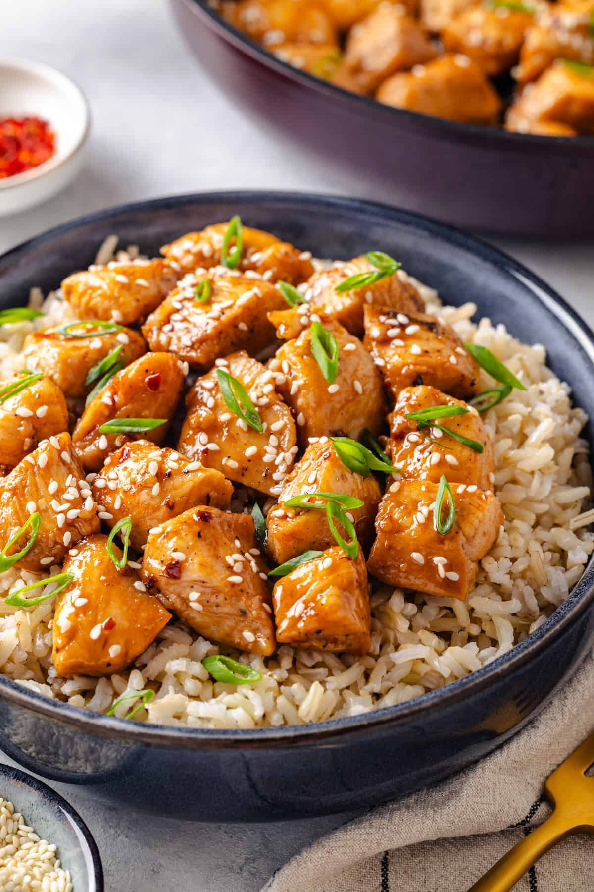 Honey garlic chicken pieces served over steamed rice with sesame seeds and sliced green onions sprinkled on top and the skillet of chicken in the background.