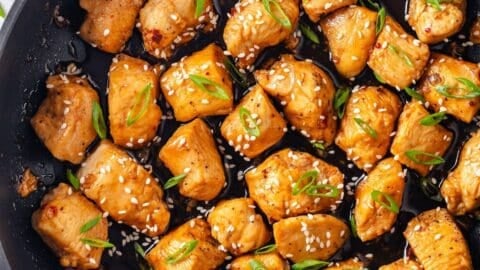 Close up of a dark skillet with a saucy honey garlic chicken dish with sesame seeds and sliced green onions sprinkled on top.