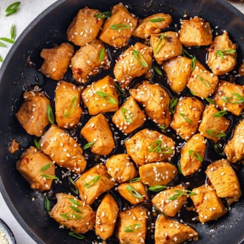 Close up of a dark skillet with a saucy honey garlic chicken dish with sesame seeds and sliced green onions sprinkled on top.