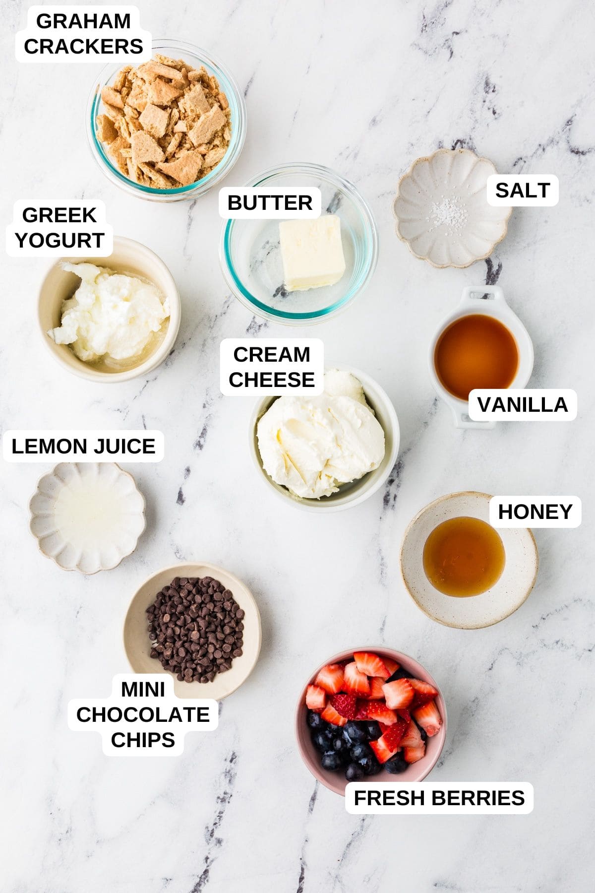 Ingredients laid out in separate bowls for making a cheesecake recipe, including graham crackers, Greek yogurt, cream cheese and fresh berries.