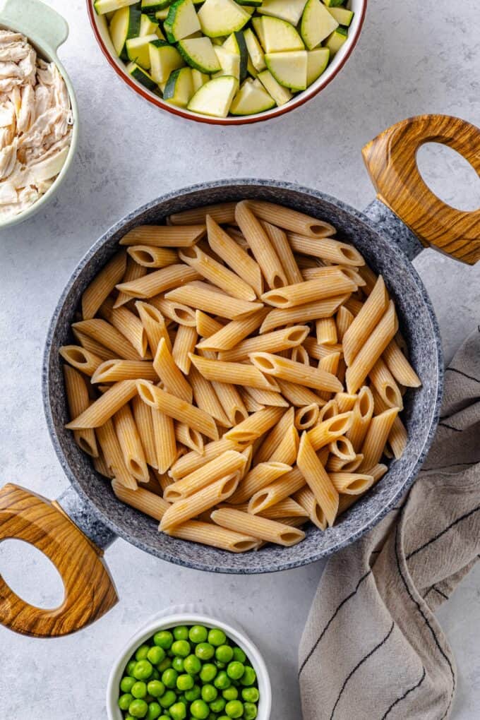 A bowl of cooked penne pasta with bowls nearby of chicken, zucchini and peas.