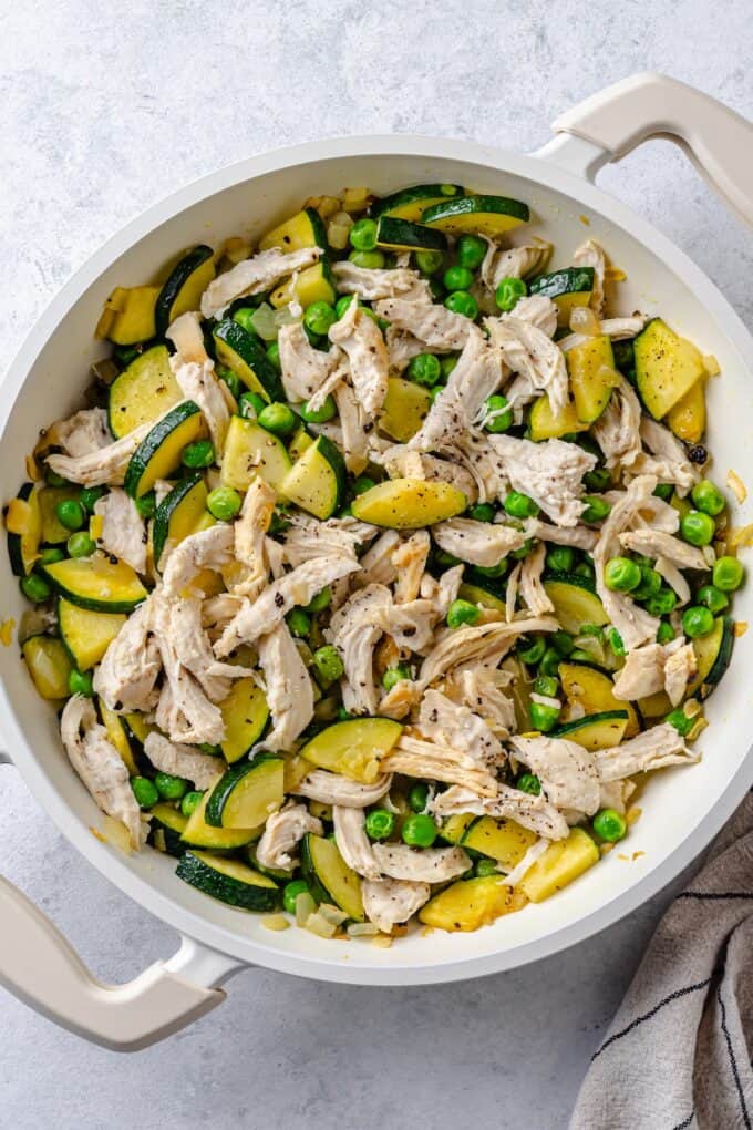 A white skillet with a mixture o zucchini, onion, green peas and cooked, shredded chicken mixed together.