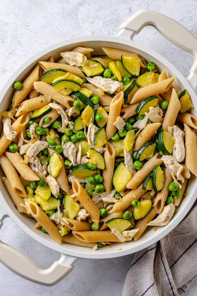 A full deep white skillet with a pasta mixture with chicken, zucchini and peas.