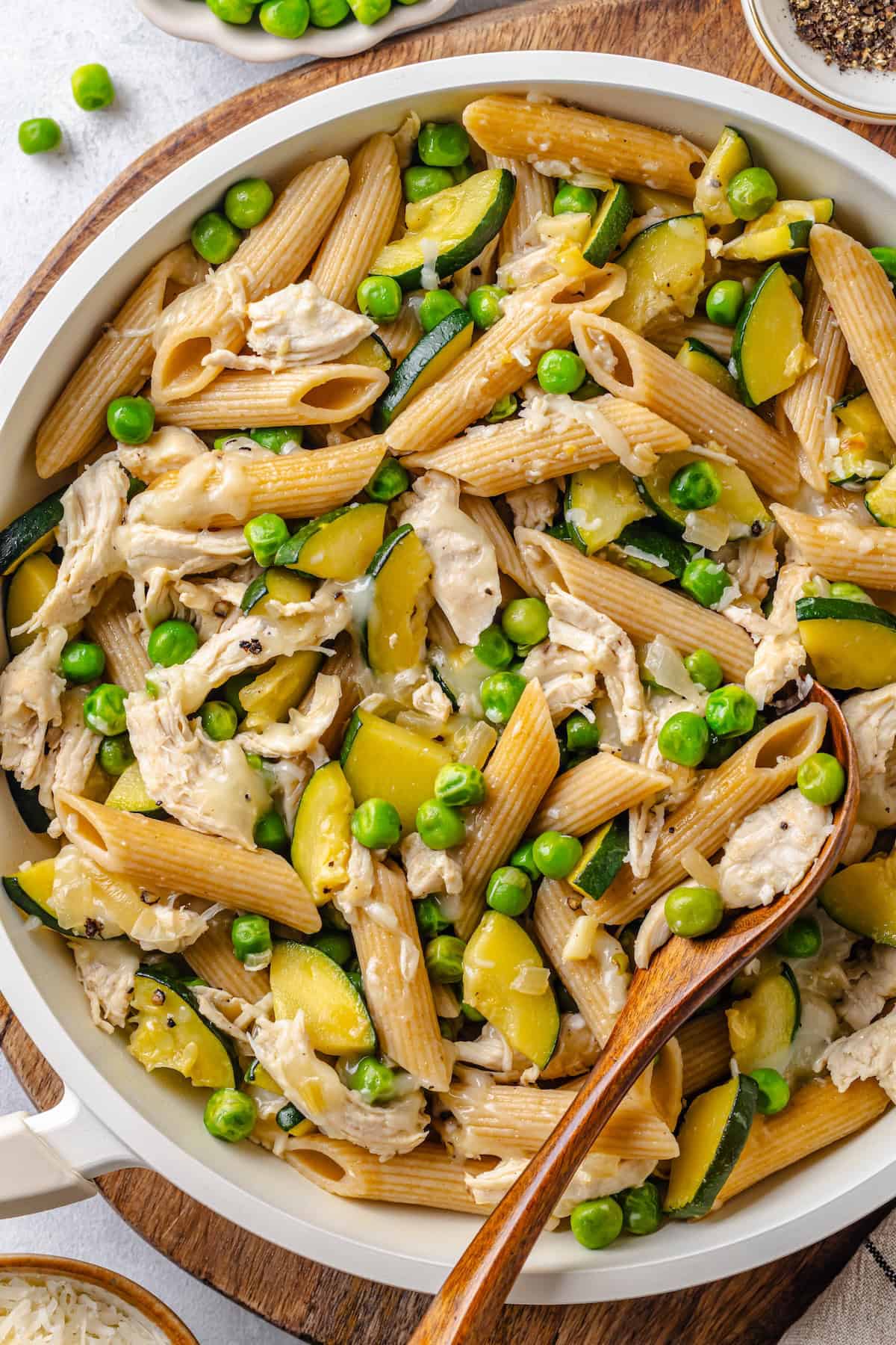 A small wooden serving spoon resting in a white skillet full of a cheesy healthy chicken pasta with zucchini and peas.