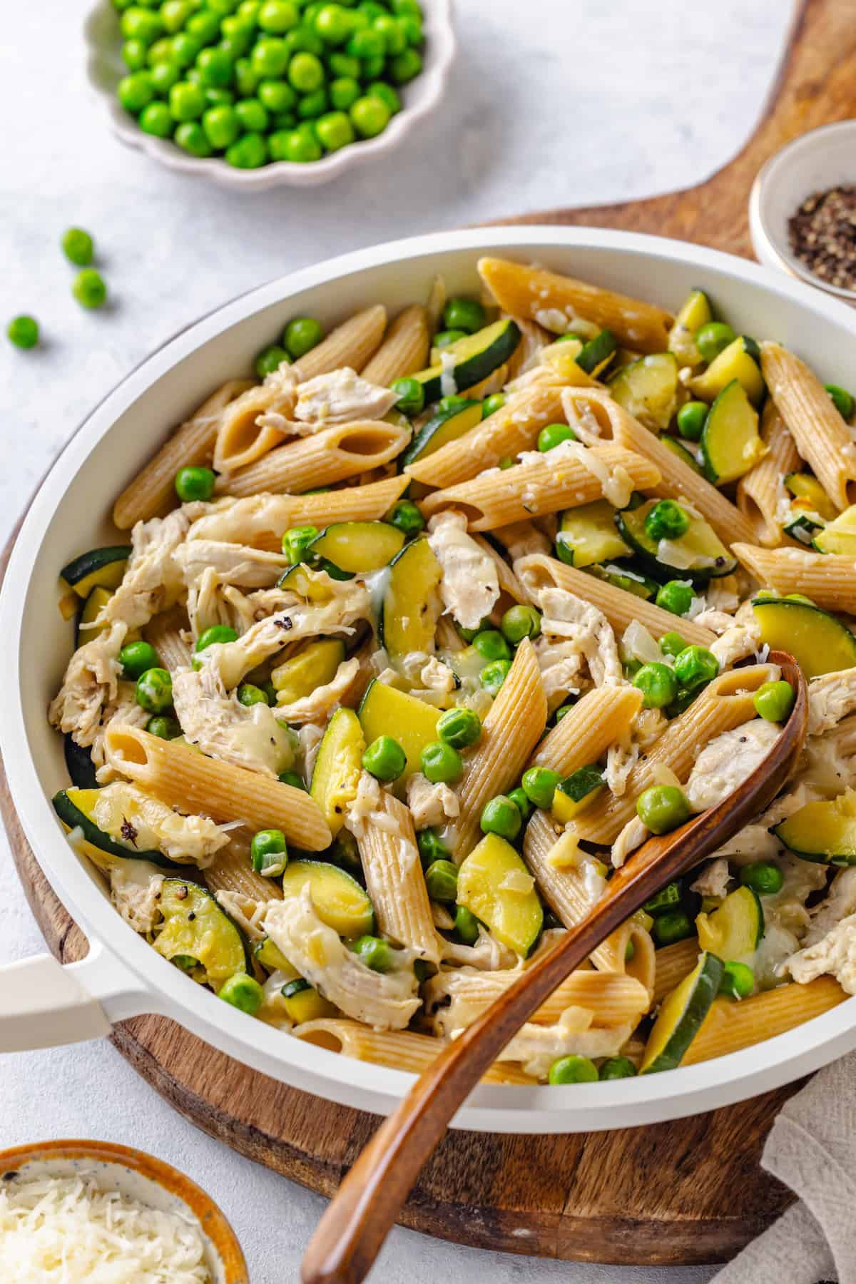 A small wooden serving spoon resting in a white skillet full of a cheesy healthy chicken pasta with zucchini and peas set on a cutting board.