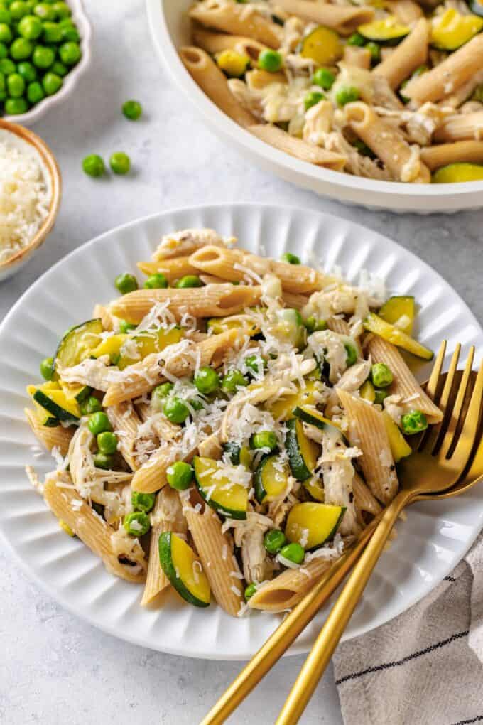 A gold fork and spoon resting on the side of a white plate filled with a chicken veggie pasta mix with Parmesan cheese on top.