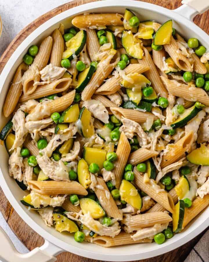 Close up of a white pan filled with a chicken pasta with vegetables set on a wooden cutting board.