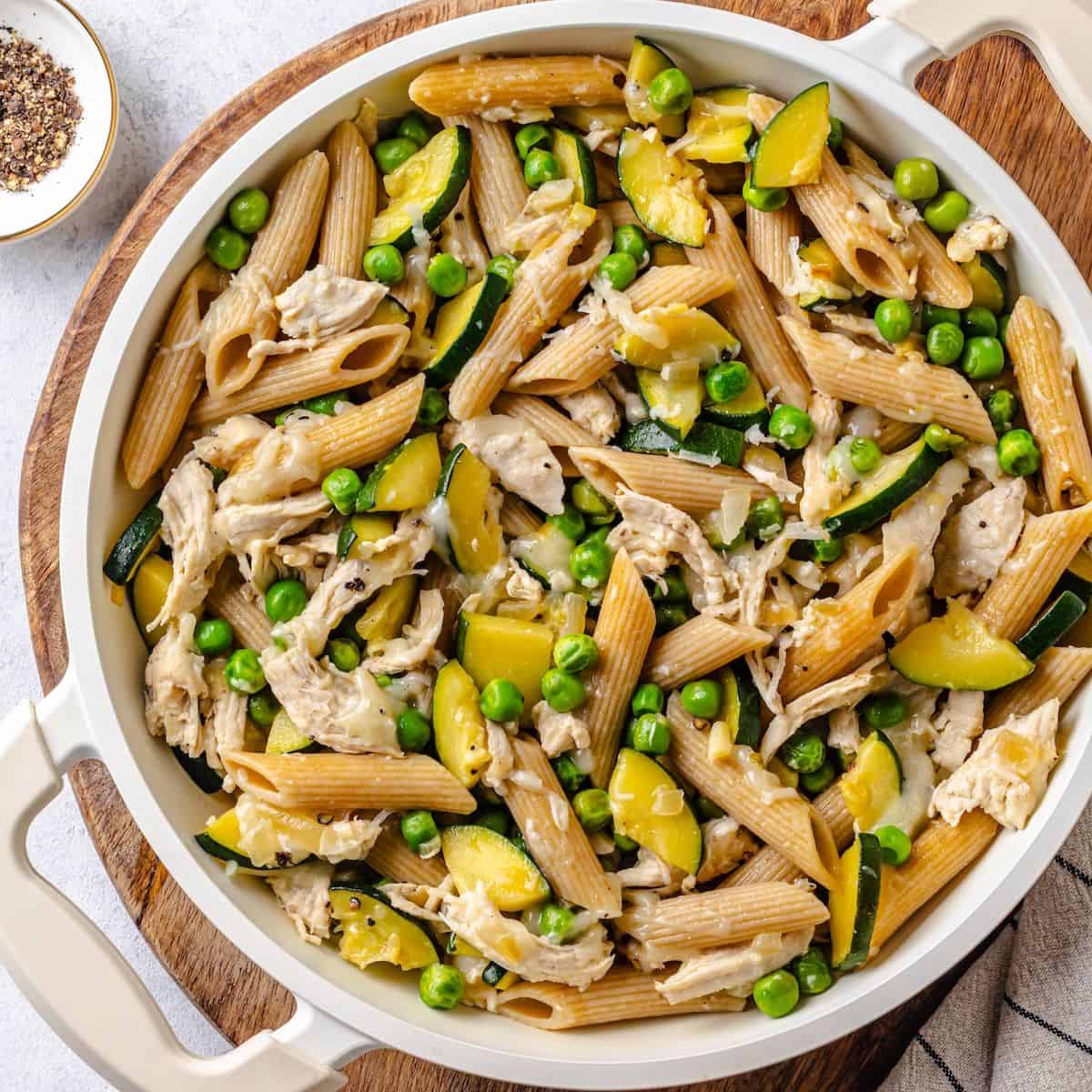 Close up of a white pan filled with a chicken pasta with vegetables set on a wooden cutting board.