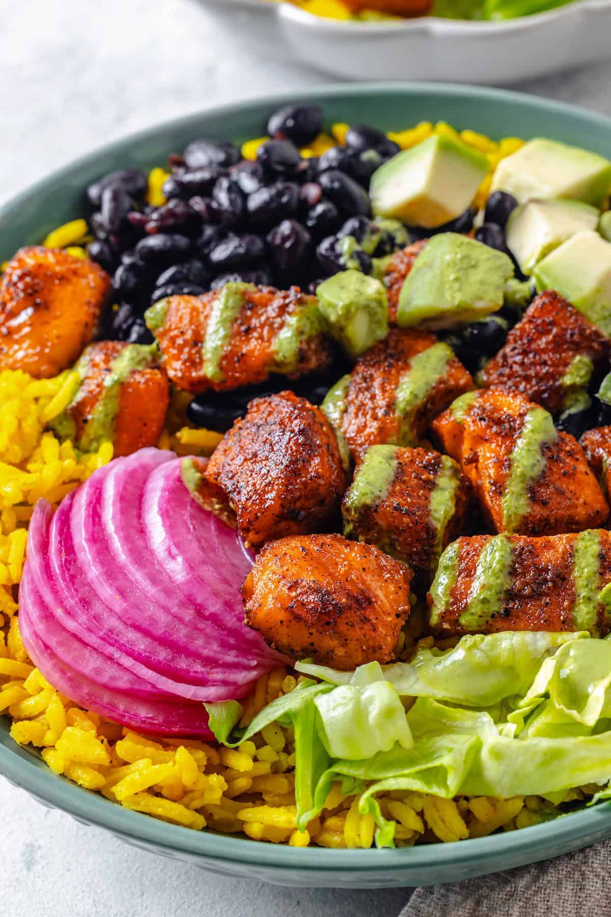 Close up of a green bowl with yellow rice topped with piles of black beans, salmon bites, chopped lettuce, pickled red onion, diced avocado and cilantro dressing drizzled on top.