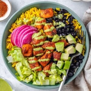 A fork resting in a green bowl with yellow rice topped with piles of black beans, salmon bites, chopped lettuce, pickled red onion, diced avocado and cilantro dressing drizzled lightly on top.
