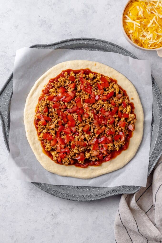 Pizza sauce spread out on a rolled out pizza dough on a pizza pan and topped with seasoned cooked taco meat and chopped tomatoes.