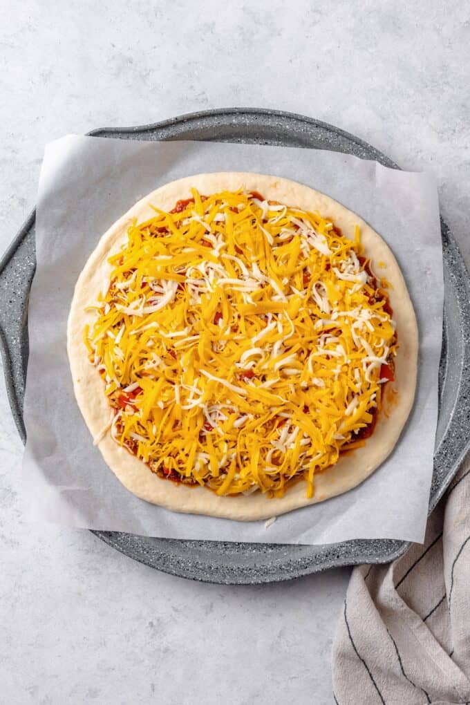 Pizza sauce spread out on a rolled out pizza dough on a pizza pan and topped with seasoned cooked taco meat. chopped tomatoes and shredded cheddar cheese.