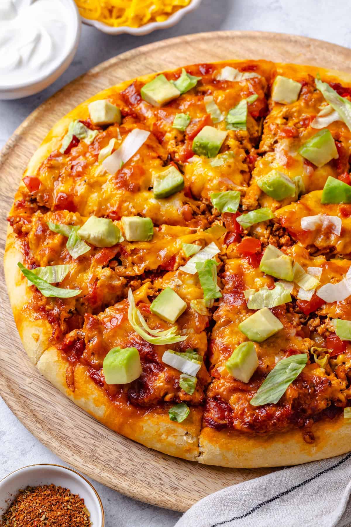 A sliced taco pizza with lettuce and avocado on top on a wooden cutting board.