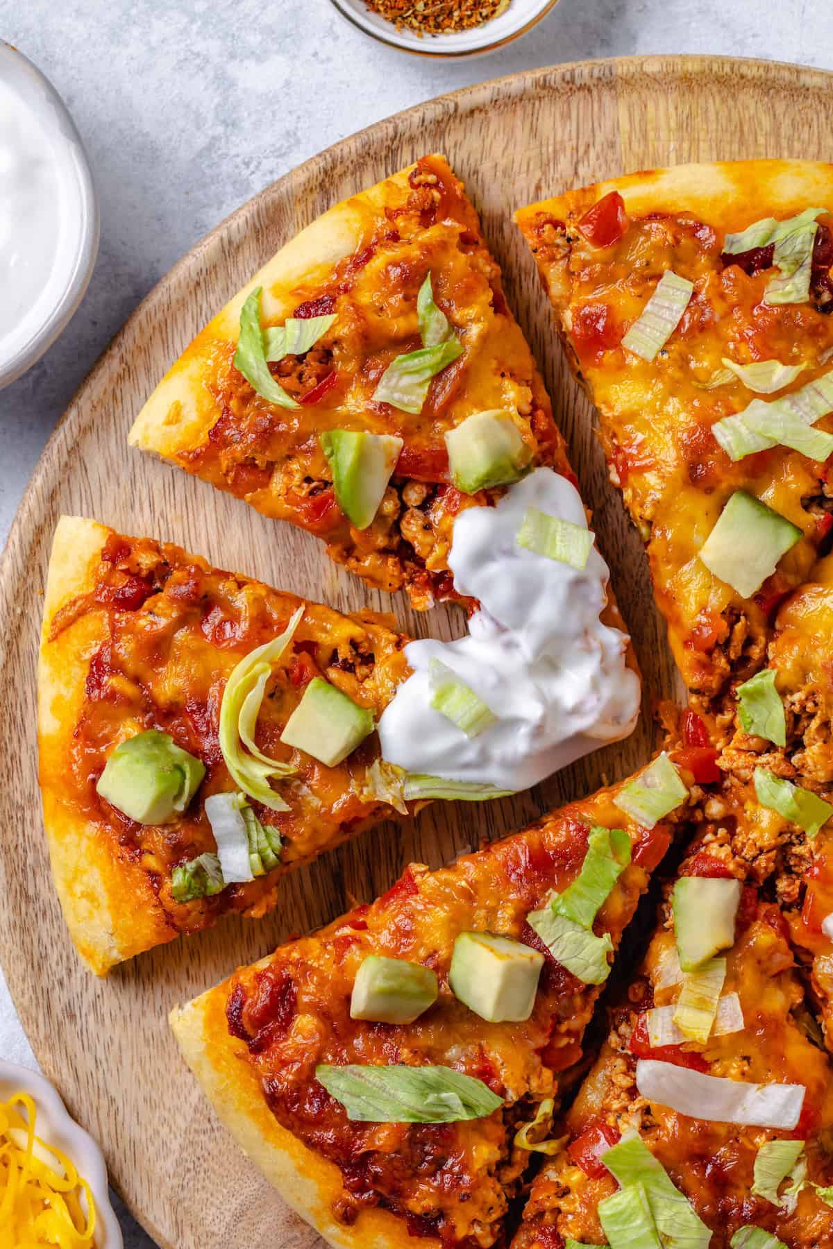 Sliced taco pizza with lettuce and avocado on top on a wooden cutting board, with sour cream dipped onto two of the slices.