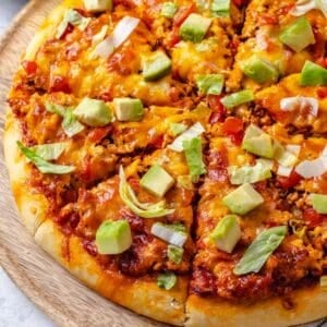 Close up of sliced homemade taco style pizza with lettuce and avocado on top on a wooden cutting board.