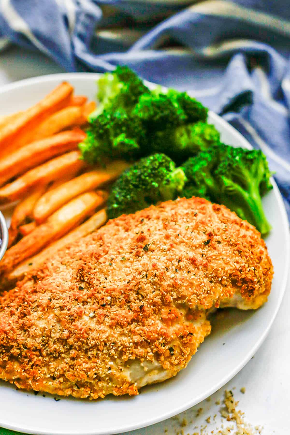 A Ranch seasoned and breadcrumb coated baked chicken breast on a white plate with sweet potato fries and broccoli.