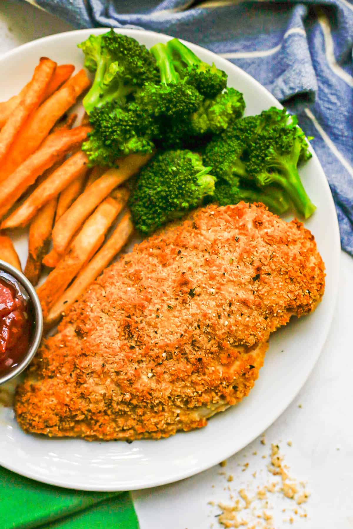 A crunchy baked chicken breast on a white plate with broccoli and sweet potato fries.