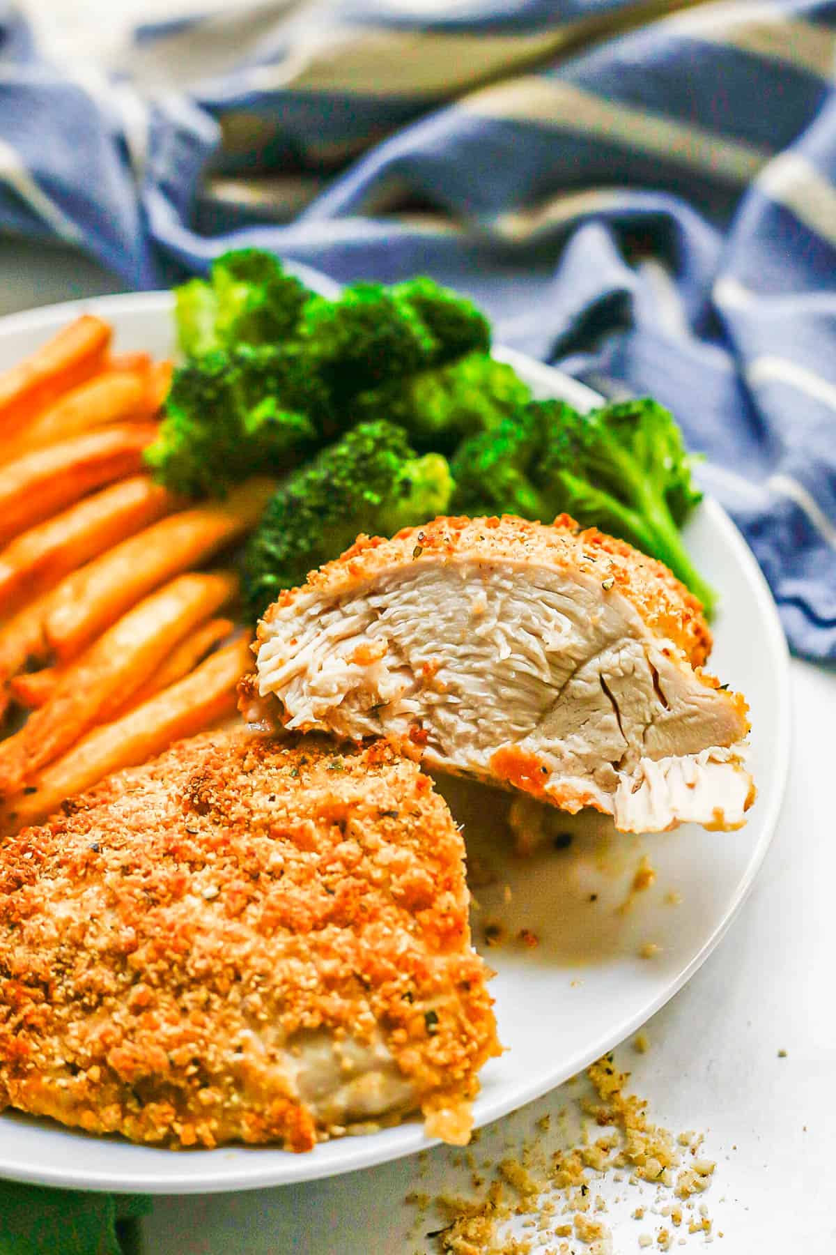 A sliced Ranch seasoned and breadcrumb coated baked chicken breast on a white plate with sweet potato fries and broccoli.