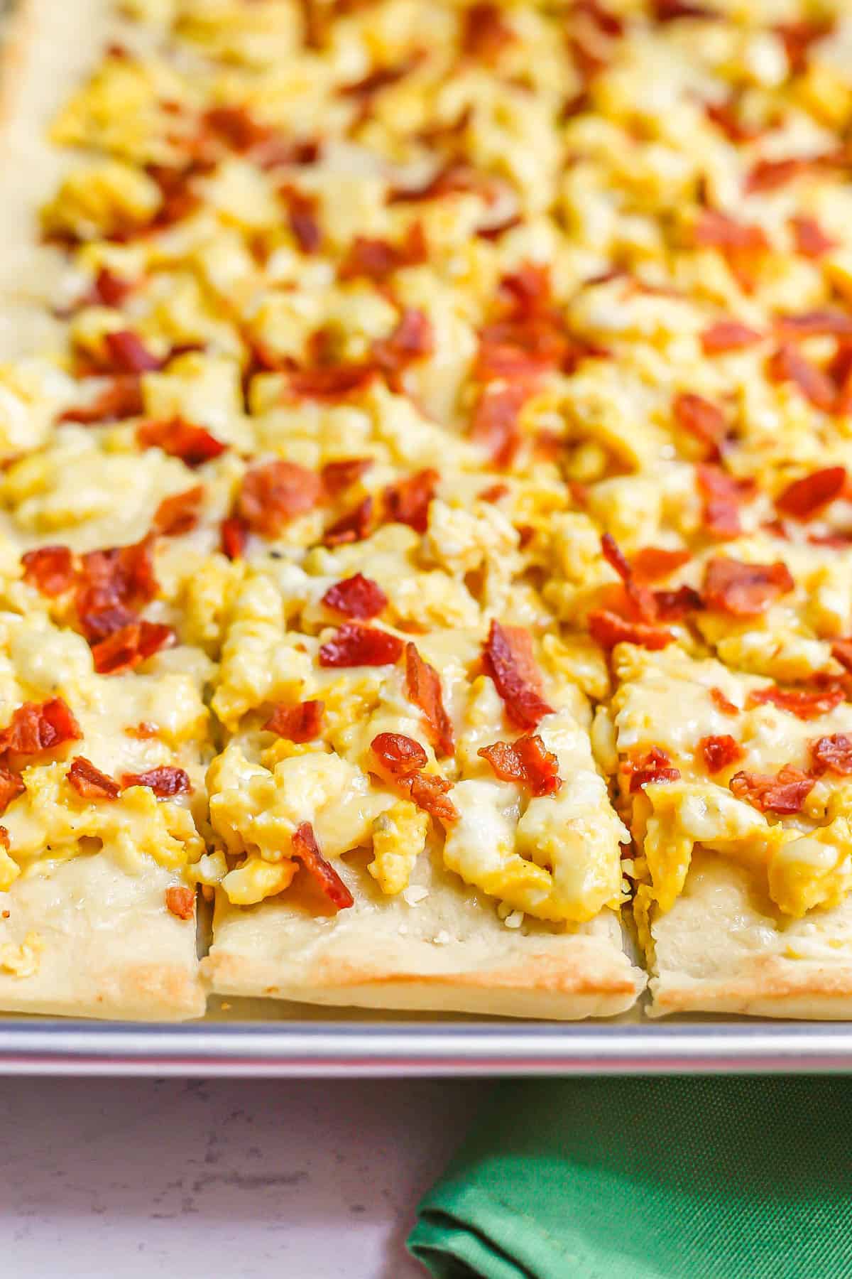 Close up of a sheet pan with a breakfast pizza with scrambled eggs, cheese and bacon cut into small squares.