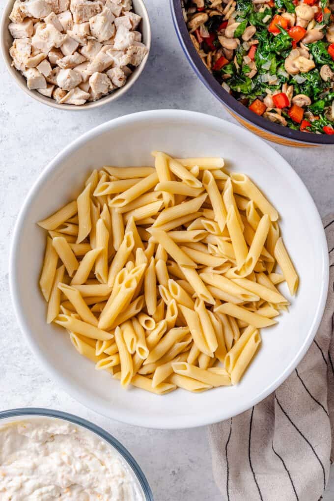 A large white bowl with cooked penne pasta, with bowls of cooked chicken, sautéed veggies and a cottage cheese mixture nearby.