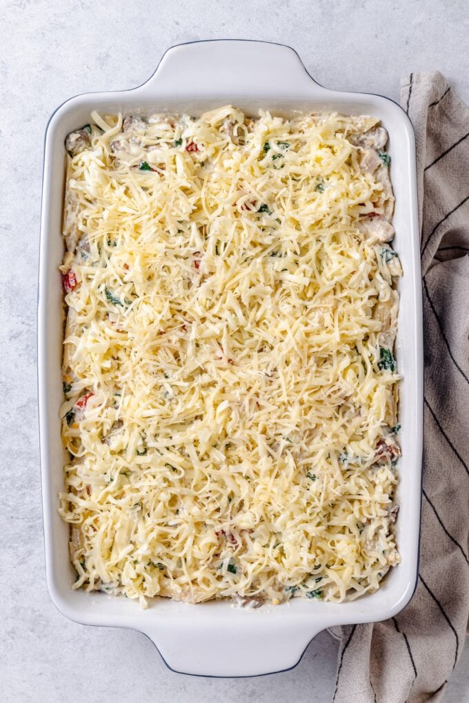 A chicken and pasta casserole topped with shredded cheese before being baked in the oven.