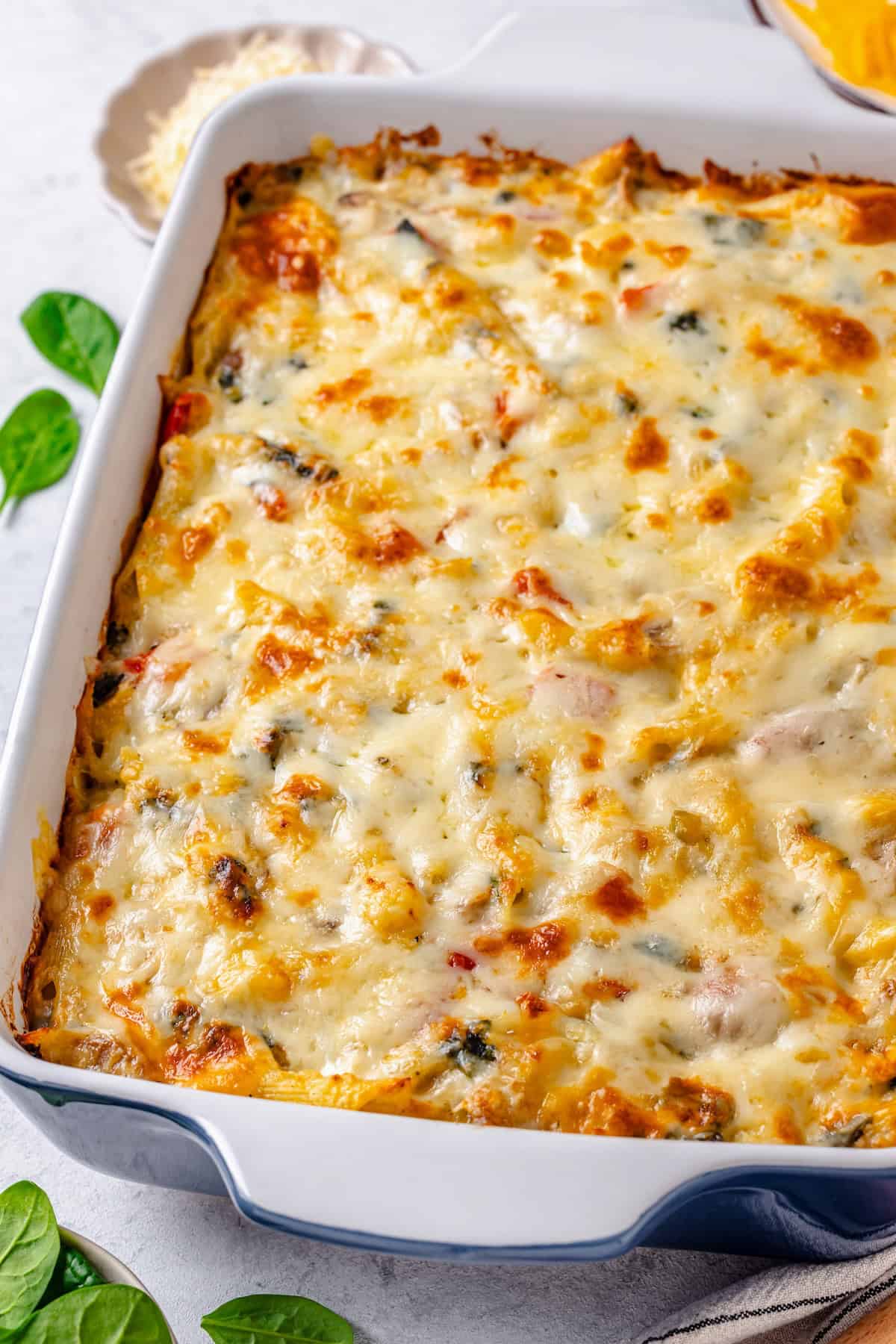 A cheesy baked casserole with chicken, pasta and veggies in a blue rectangular casserole dish with baby spinach sprinkled on the side.