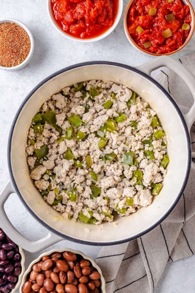 A large white pot with sautéed onion, garlic and green pepper with cooked ground turkey and bowls of tomatoes and beans and seasoning nearby.