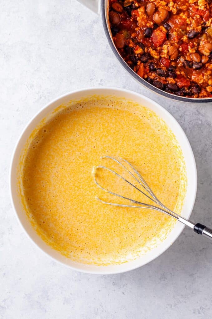 A whisk resting in a white bowl with a cornbread batter in it with a pot of chili to the side.