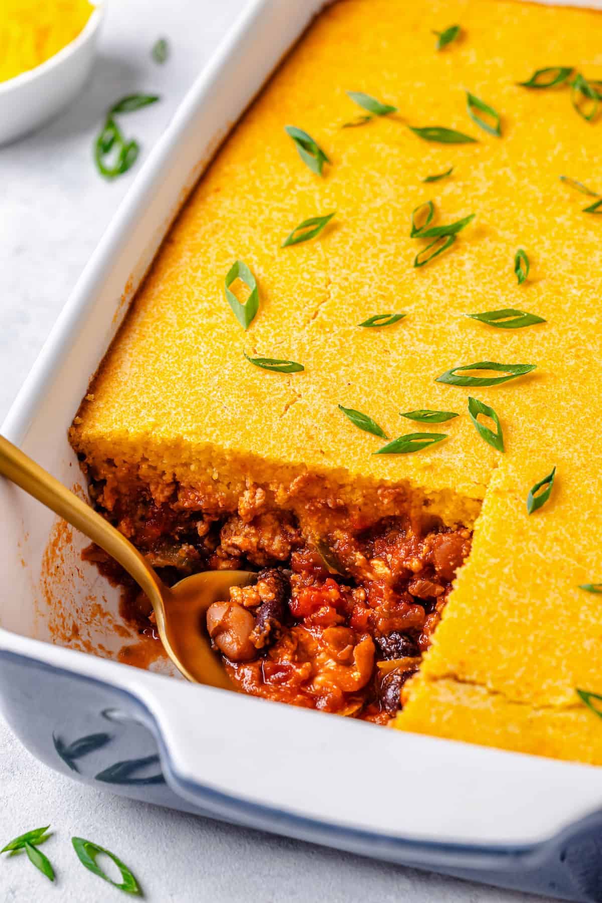 A spoon resting in the corner of a casserole dish with a square piece of chili and cornbread cut out of it.