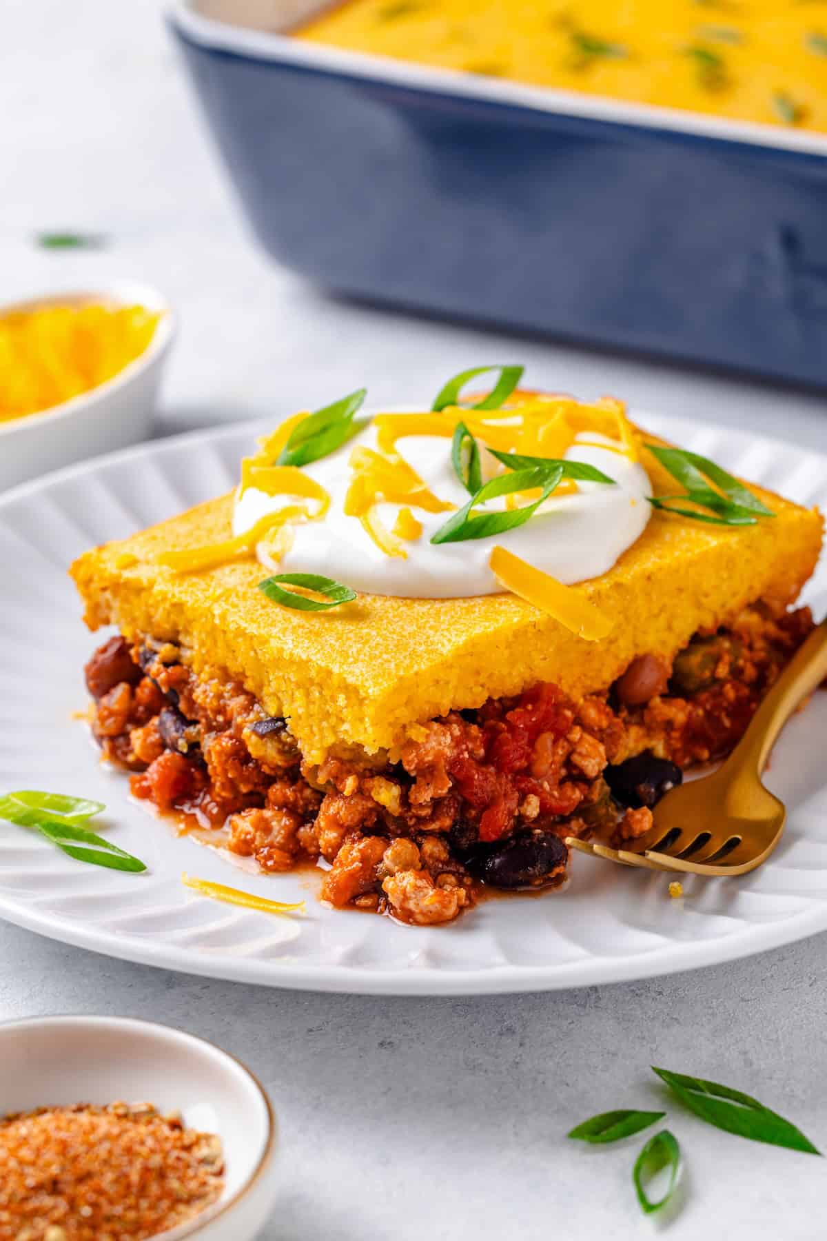 A serving of a cornbread topped chili casserole on a white plate with a fork to the side and sour cream, cheese and sliced green onions on top.