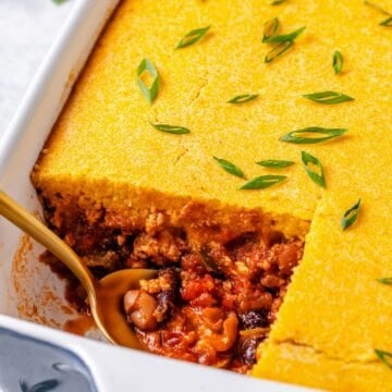 Close up of a spoon resting in the corner of a casserole dish with a square piece of chili and cornbread cut out of it.