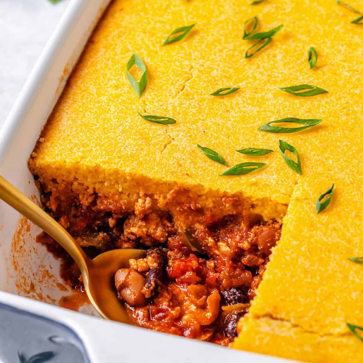 Close up of a spoon resting in the corner of a casserole dish with a square piece of chili and cornbread cut out of it.