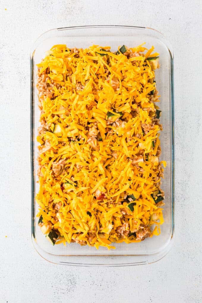 A glass casserole dish with a chicken, rice and veggie mix spread out with shredded cheddar cheese on top.