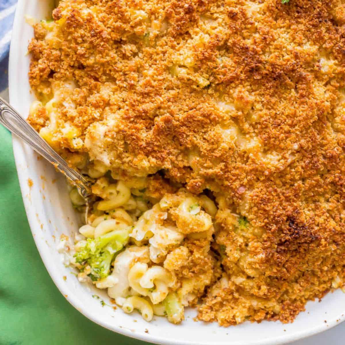 Close up of a silver serving spoon resting in a white casserole dish with a chicken and broccoli mac and cheese topped with breadcrumbs.