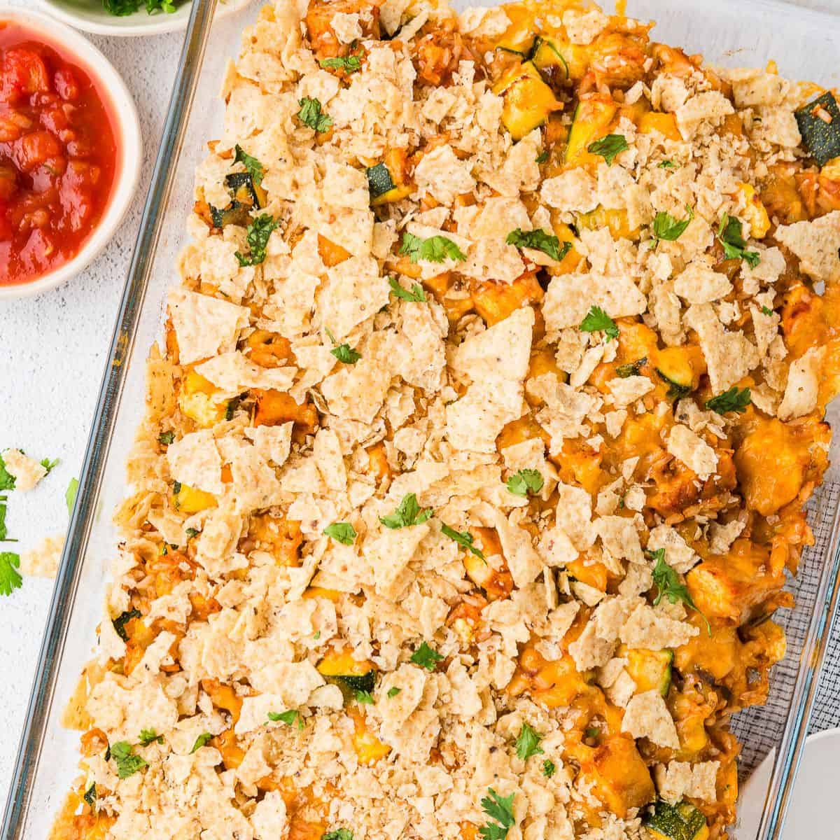 Close up of a baked chicken taco casserole with cheese and tortilla chips on top in a glass baking dish with sprinkled cilantro on top and a bowl of salsa to the side.