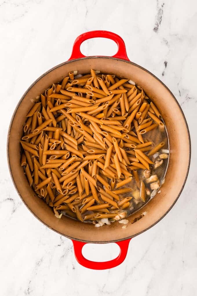 A large red pot with penne pasta added on top of some mushrooms and onion.