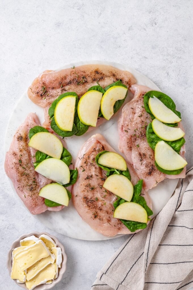 Butterflied and seasoned chicken breasts on a plate with baby spinach and sliced apples on one side of each chicken breast.