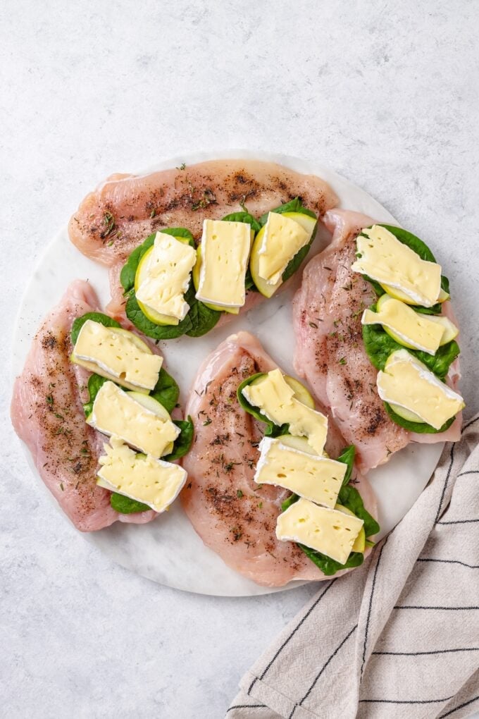 Butterflied and seasoned chicken breasts on a plate with baby spinach, sliced apples and brie cheese slices on one side of each chicken breast.